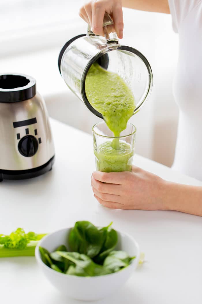 healthy eating, cooking, vegetarian food, dieting and people concept - close up of young woman with green vegetables pouring detox shake or smoothie from blender jar to glass at home